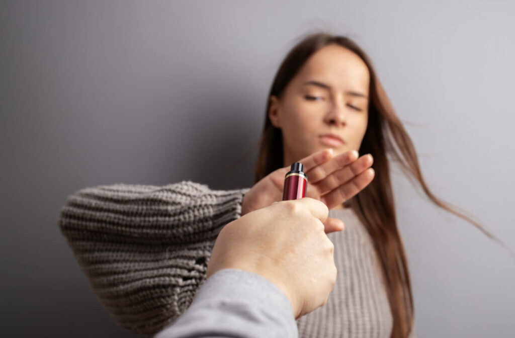 A young adult in front of a gray background raising a hand and turning down an e-cigarette vaping device.