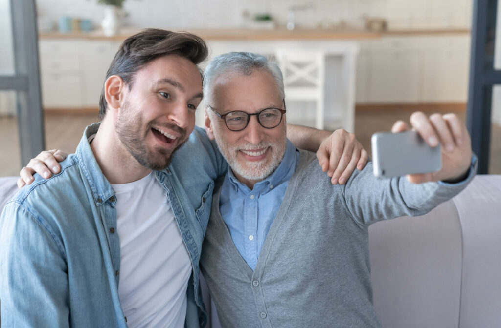 A senior and their adult child sit on a couch together, posing for a selfie. The parent and child have their arms around each other's shoulders, and are smiling broadly.